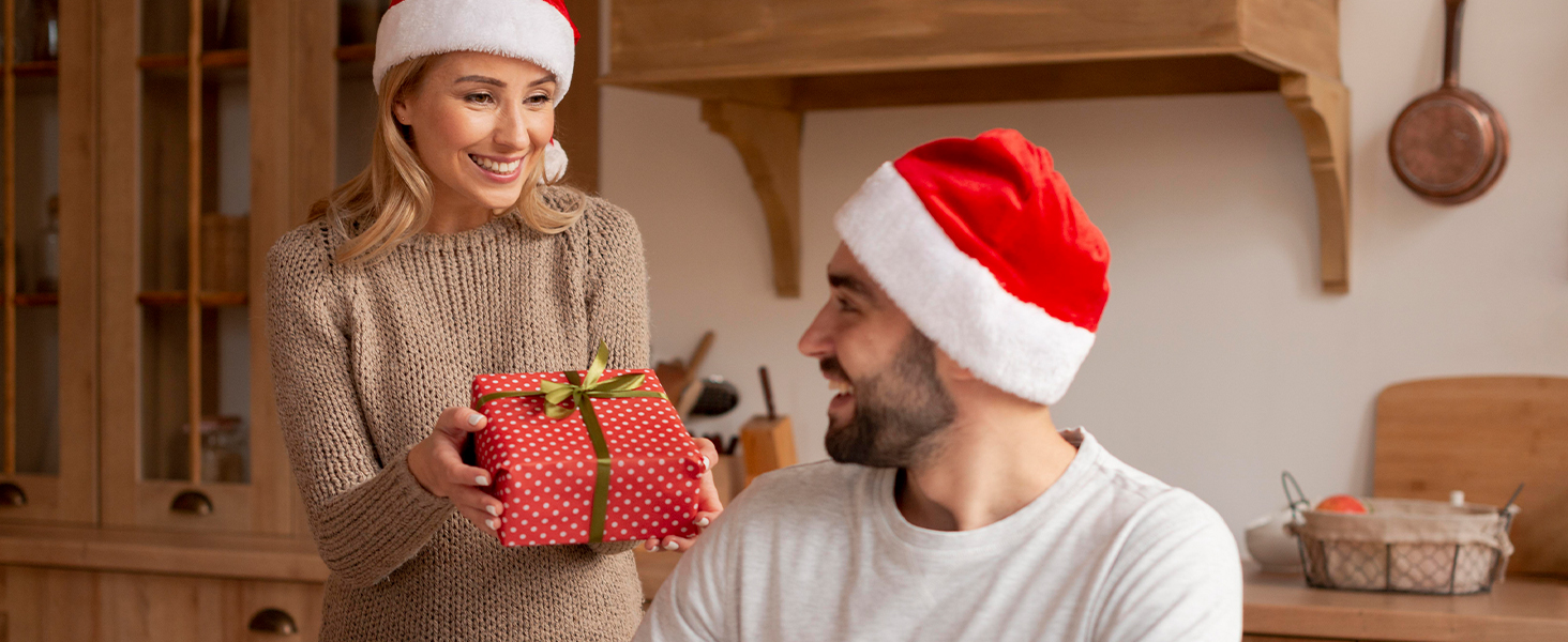 Two individuals exchanging a red wrapped gift in a cozy kitchen setting, both wearing winter attire.